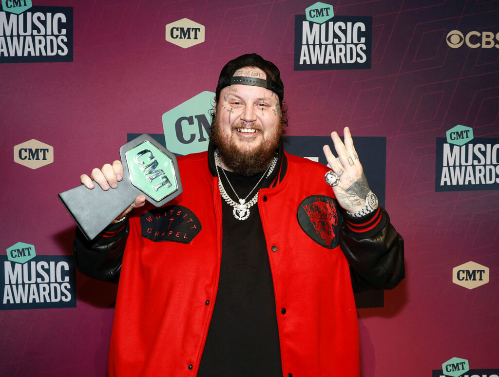 Jelly Roll on the 2023 CMT Music Awards red carpet wearing a red jacket and holding an award. 