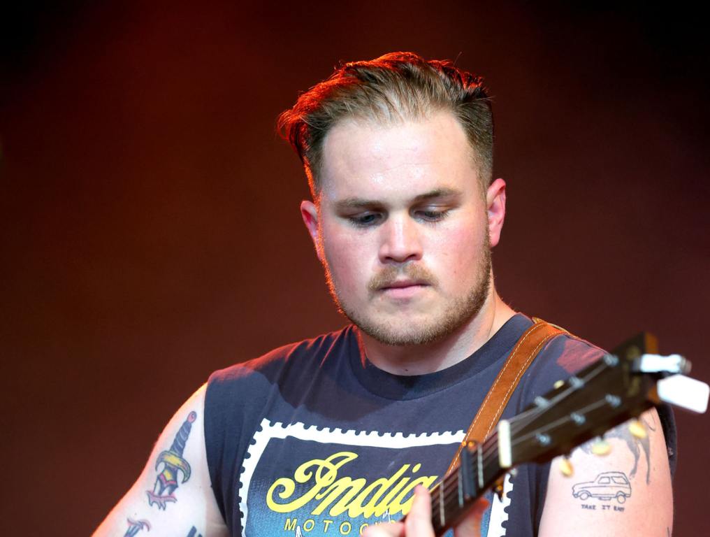 Zach Bryan looking down playing guitar in a black t-shirt