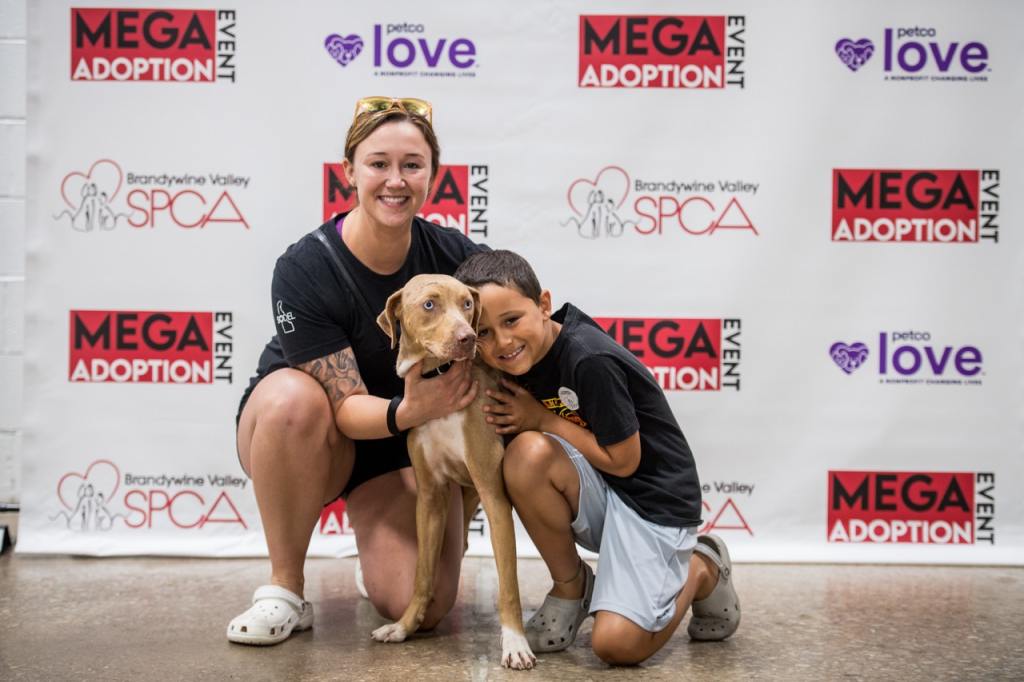 A woman and young boy kneel down to hug a medium sized light brown dog in front of a step and repeat sign with the Brandywine Valley SPCA logo and Mega Adoption Event written on it. So many families will go home with their new furry family member at the Brandywine Valley SPCA's Mega Adoption Event 2024.