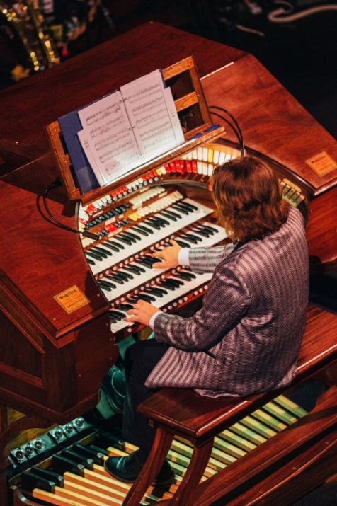 Free Movies this weekend at Colonial Theater! This photo depicts a man in a purple suit sitting at an organ in the Colonial Theater, seemingly playing along with a classic movie. 